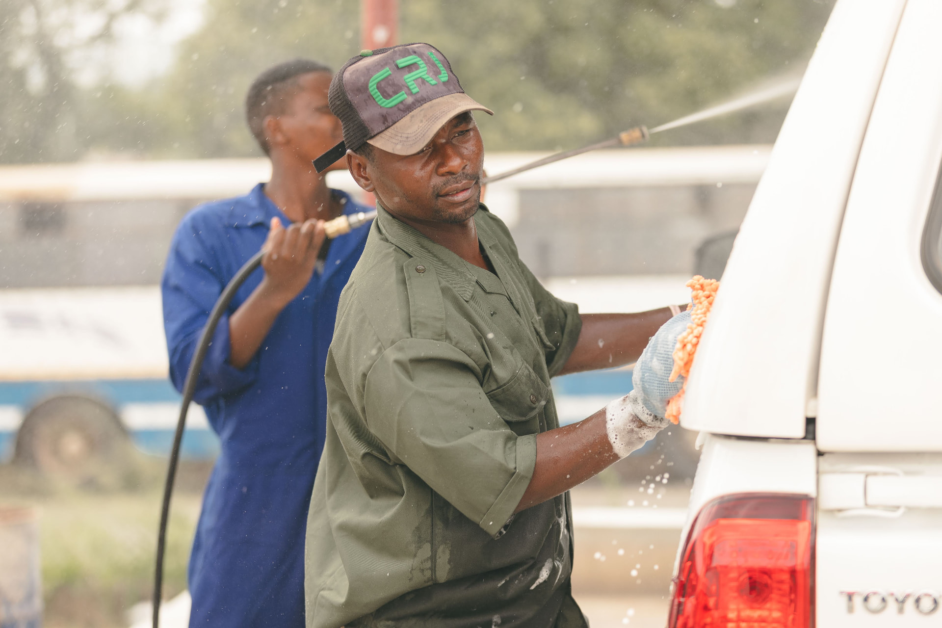 MAPS Village attendant pressure washing thick foam from a vehicle at sunset
